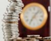 A close-up image of stacked coins with a blurred clock, symbolizing time and money relationship.